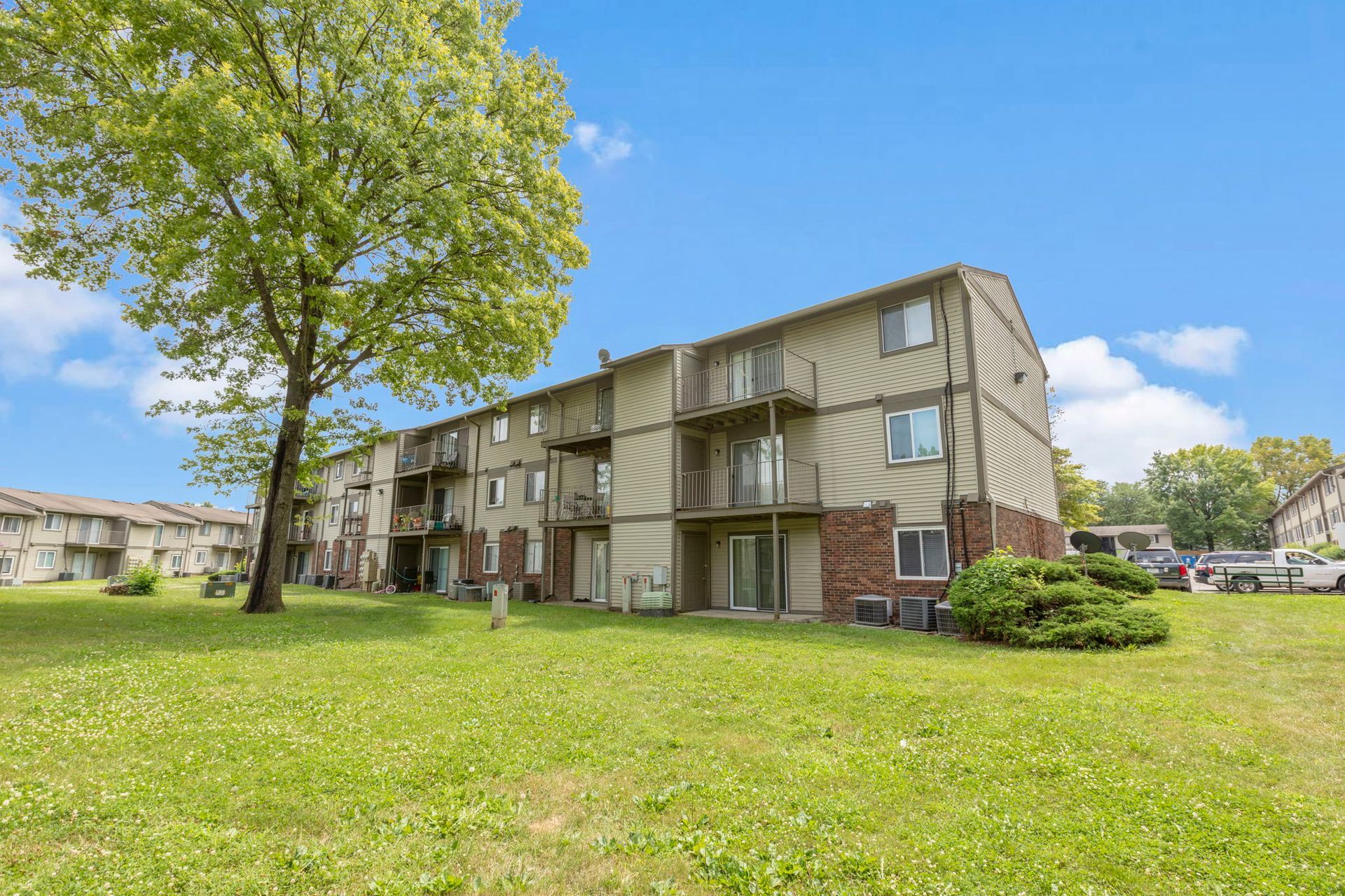 Apartment building with balconies, surrounded by green grass and a tree on a sunny day.