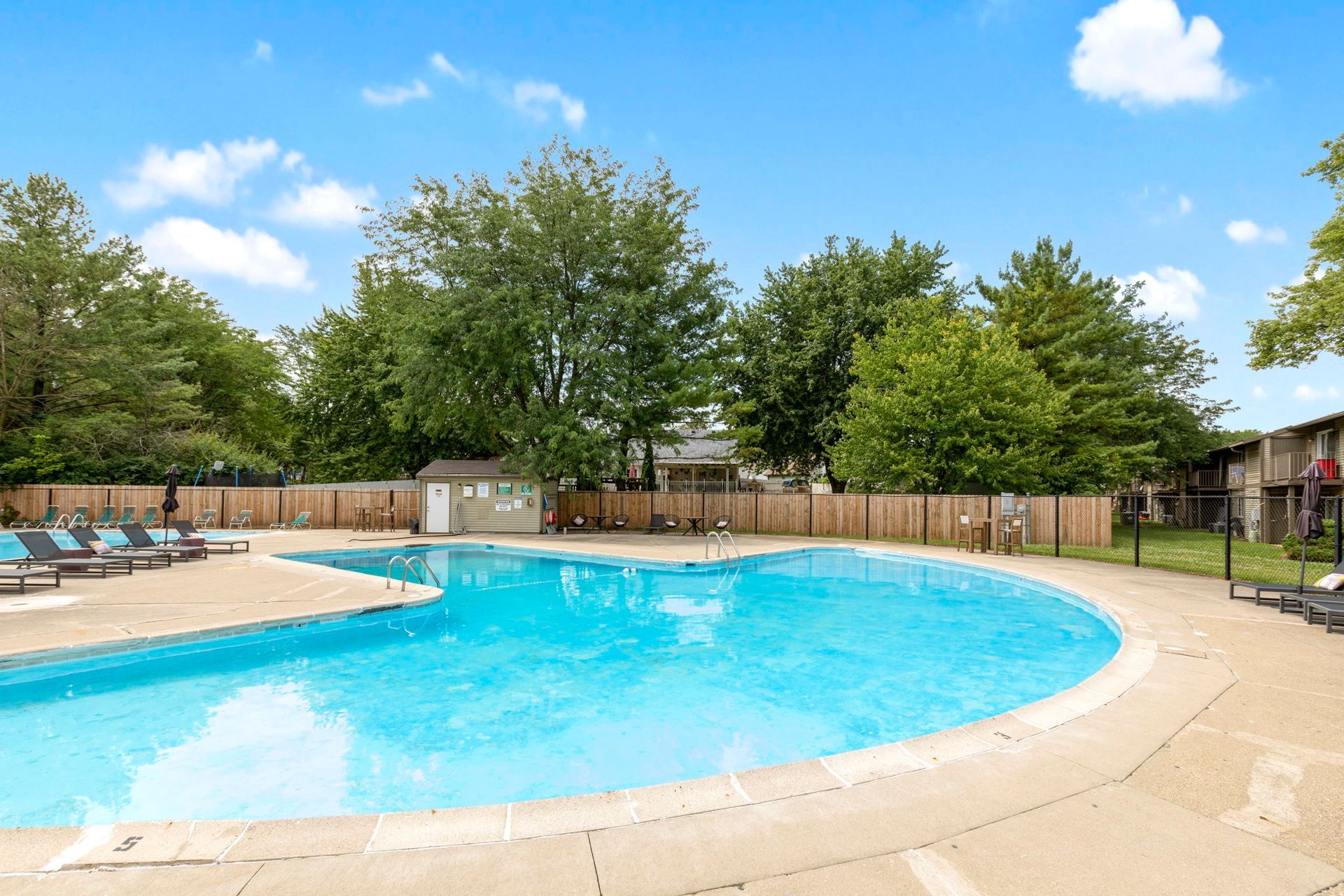 Swimming pool with blue water, surrounded by a concrete deck, fence, and trees under a cloudy sky.