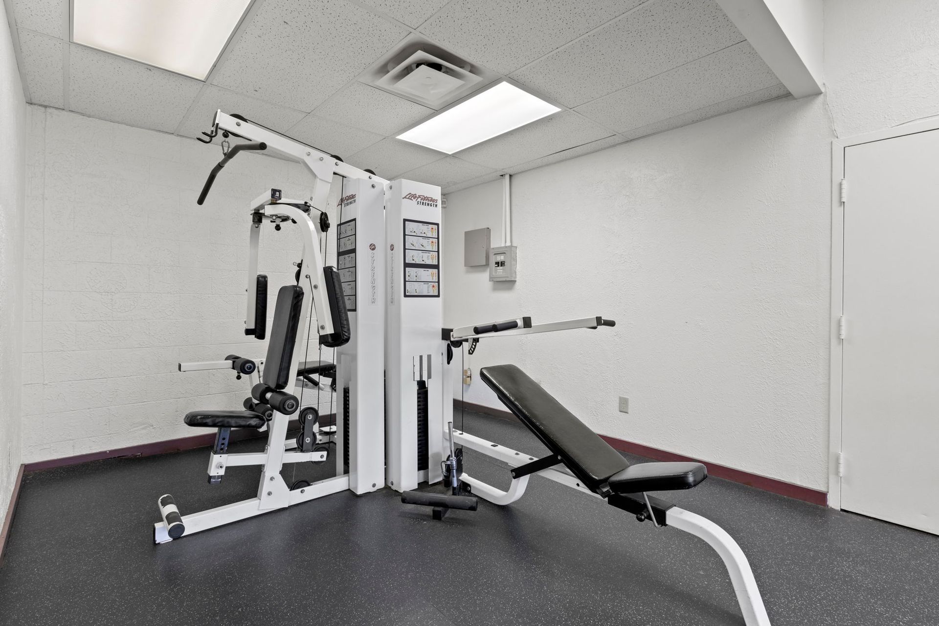 Gym with multi-station weight machine and bench on black rubber flooring. White walls and a closed door.