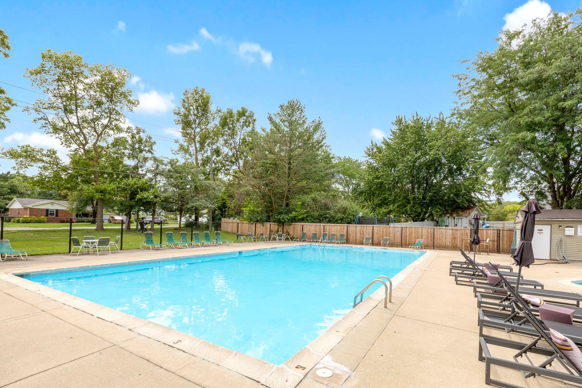 A rectangular outdoor swimming pool surrounded by trees, lounge chairs, and a wooden fence under a blue sky.