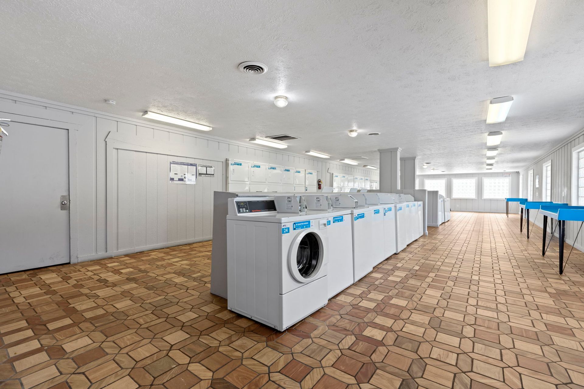 Laundry room with multiple washing machines in a row, brown tile floor, bright overhead lighting.