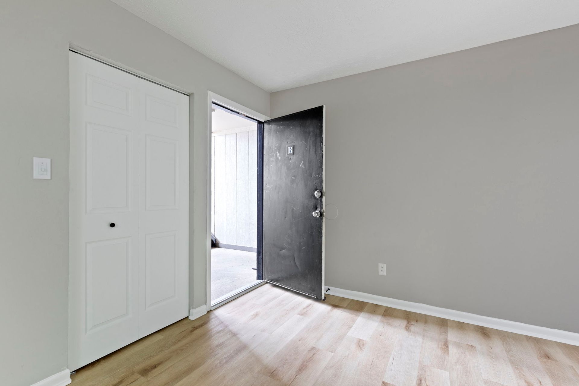 Interior view of an apartment entry. Features a white closet door, grey walls, wood flooring, and an open black door.