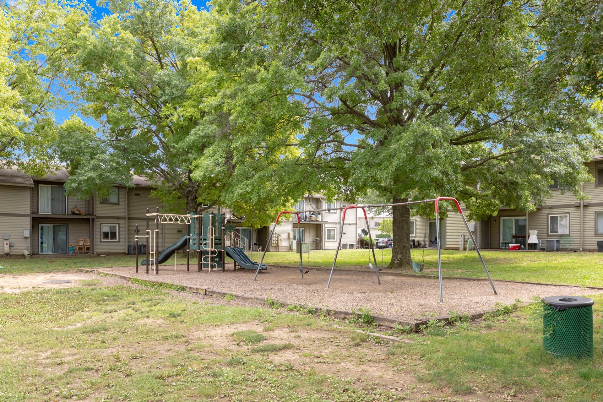 Playground in a grassy area with apartment buildings in the background; swings and slides.