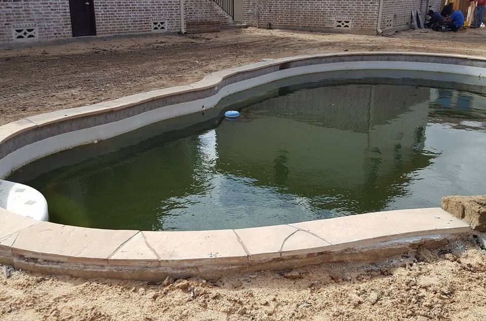 A large swimming pool is sitting in the dirt in front of a brick building.