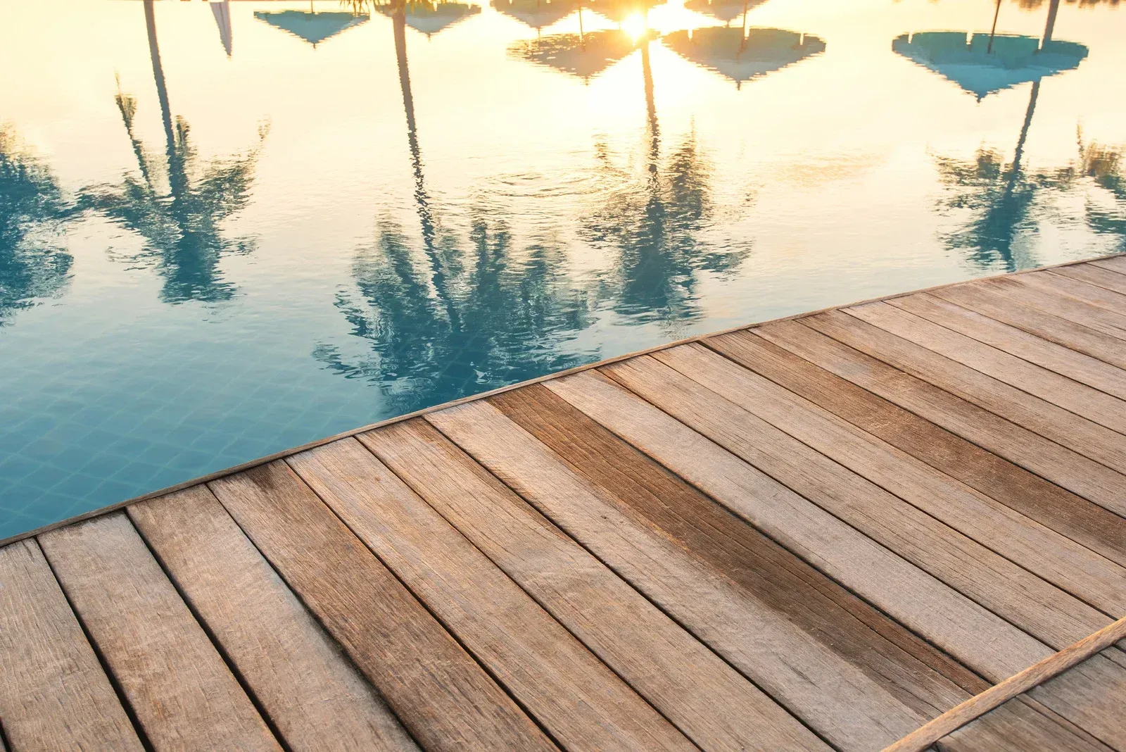 Wooden dock next to a pool reflecting palm trees and umbrellas at sunset.
