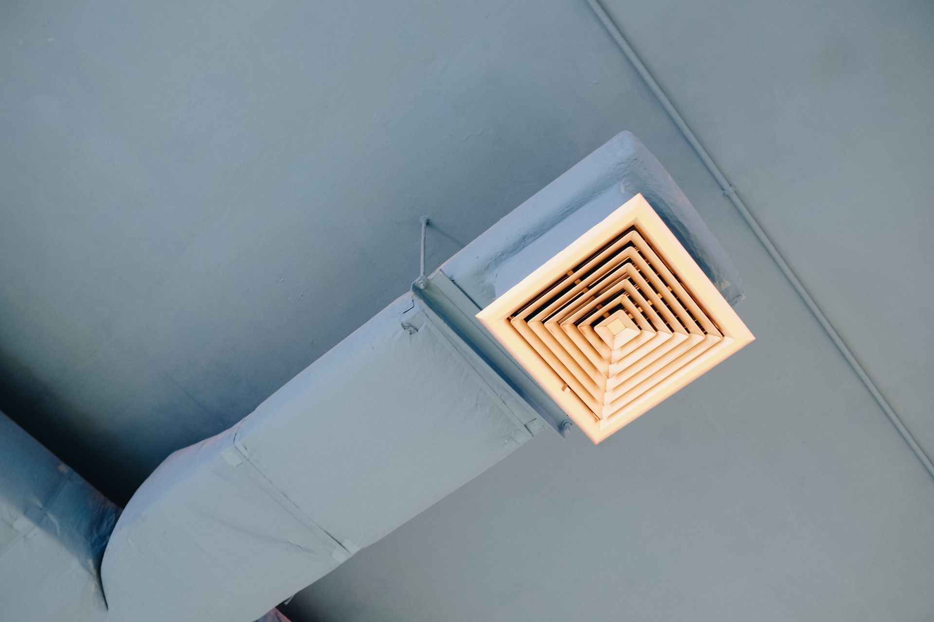 A ceiling air vent connected to a rectangular duct painted in light gray.