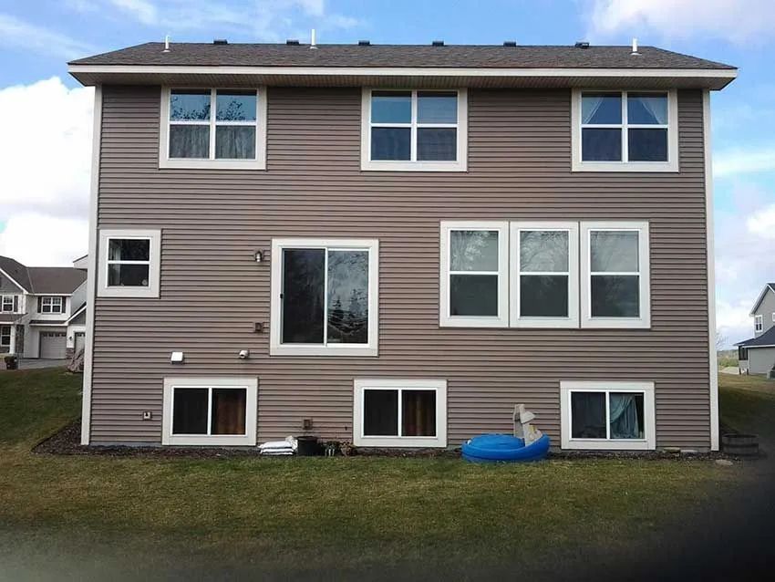 Rear view of two-story house with brown siding and multiple windows. Blue sky and green grass.