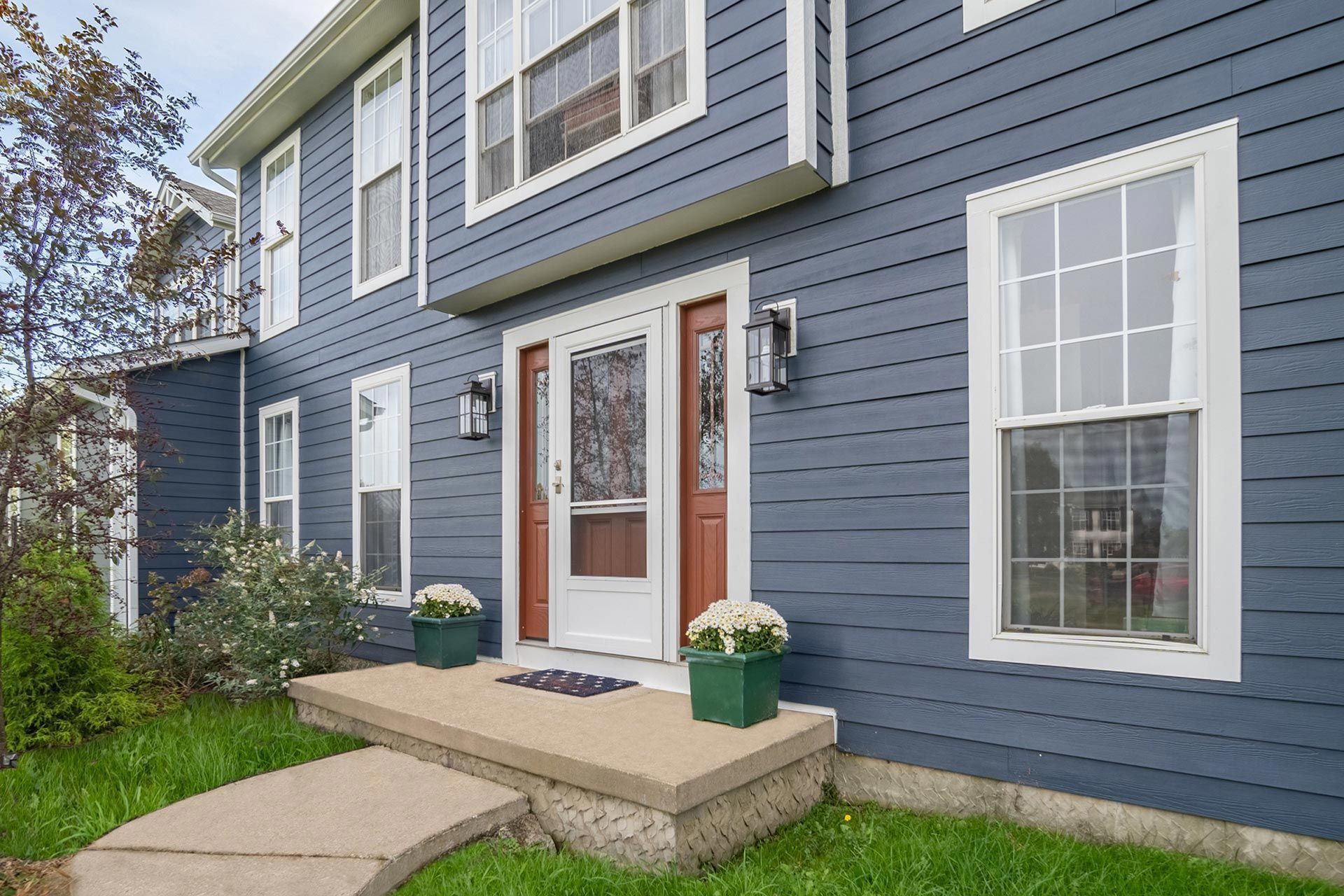 Two-story blue house with white trim, brown front door, and potted flowers on the porch.