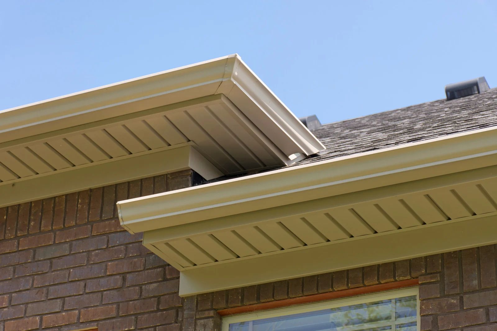 Beige soffit and gutters on a brick house with a dark shingled roof against a blue sky.