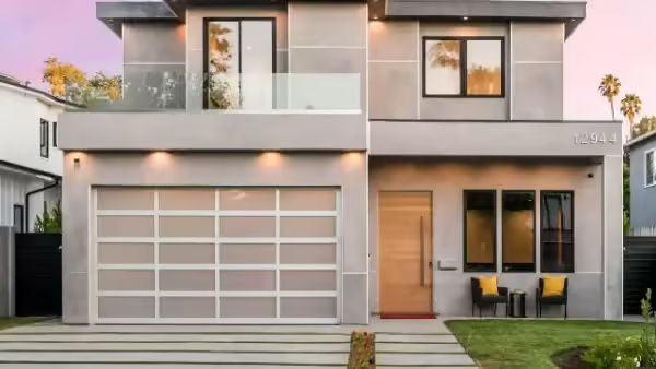 Modern gray two-story house with glass garage door, windows, and a balcony; front yard with lawn and sidewalk.