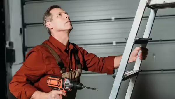 Man in red shirt and overalls using a drill while climbing a ladder, looking up in a garage.