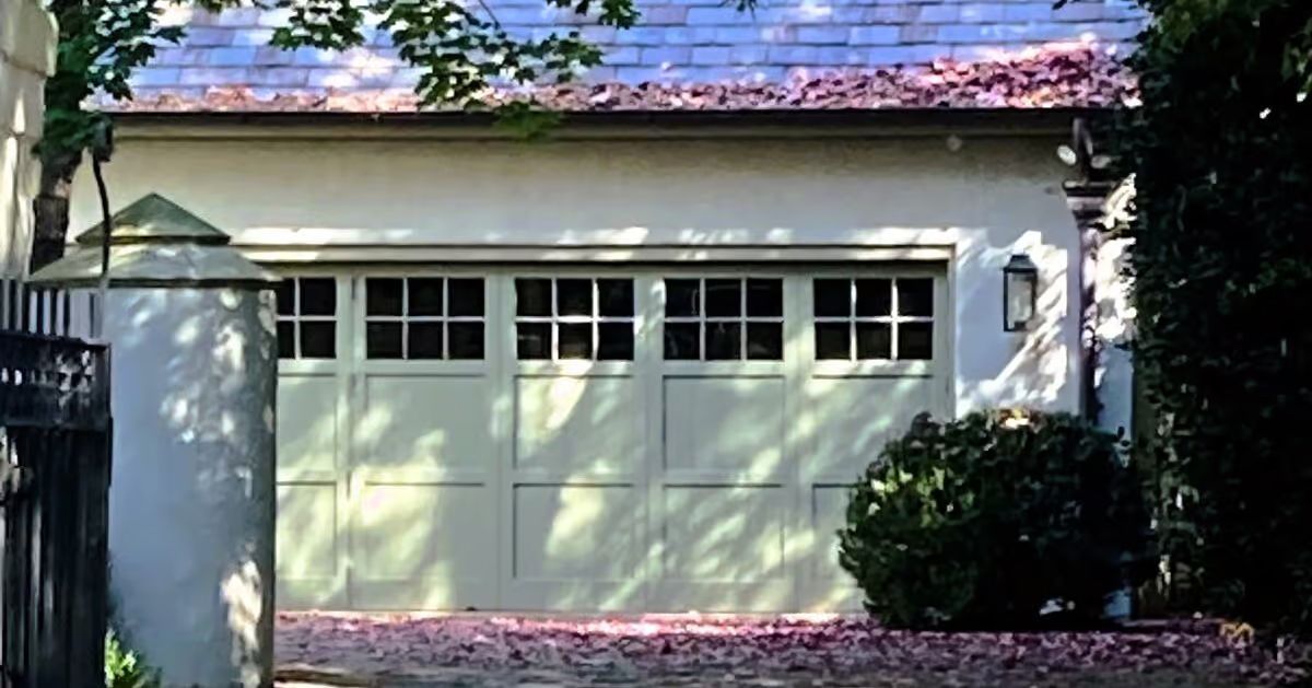 Light green garage door with multiple window panes, beige building, brick drive, and foliage.