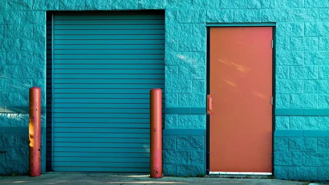 Teal building with a teal garage door and an orange door, flanked by orange poles.