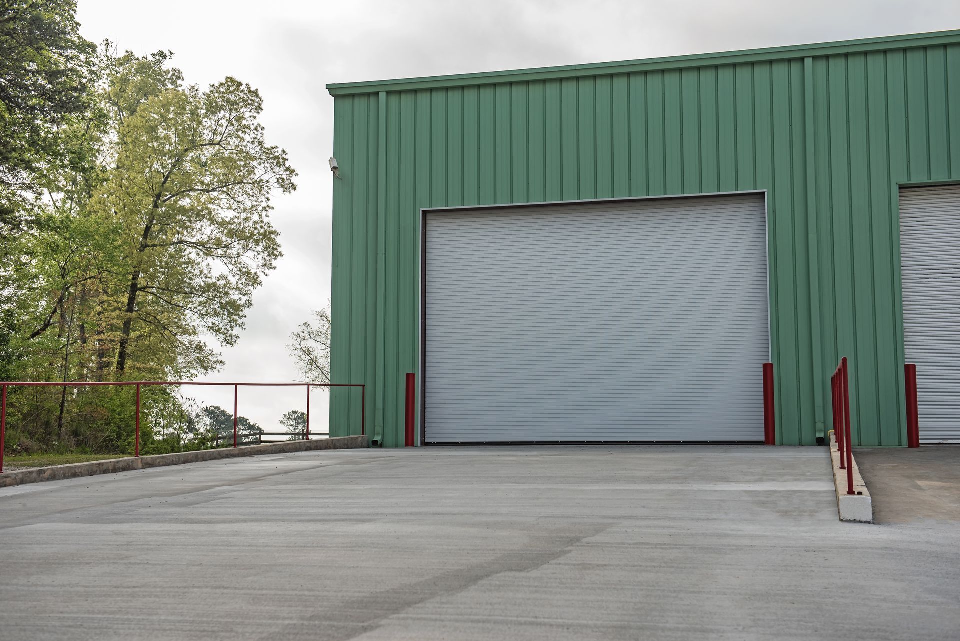 Green metal building with closed gray roll-up door. Concrete ramp and a red railing are present.