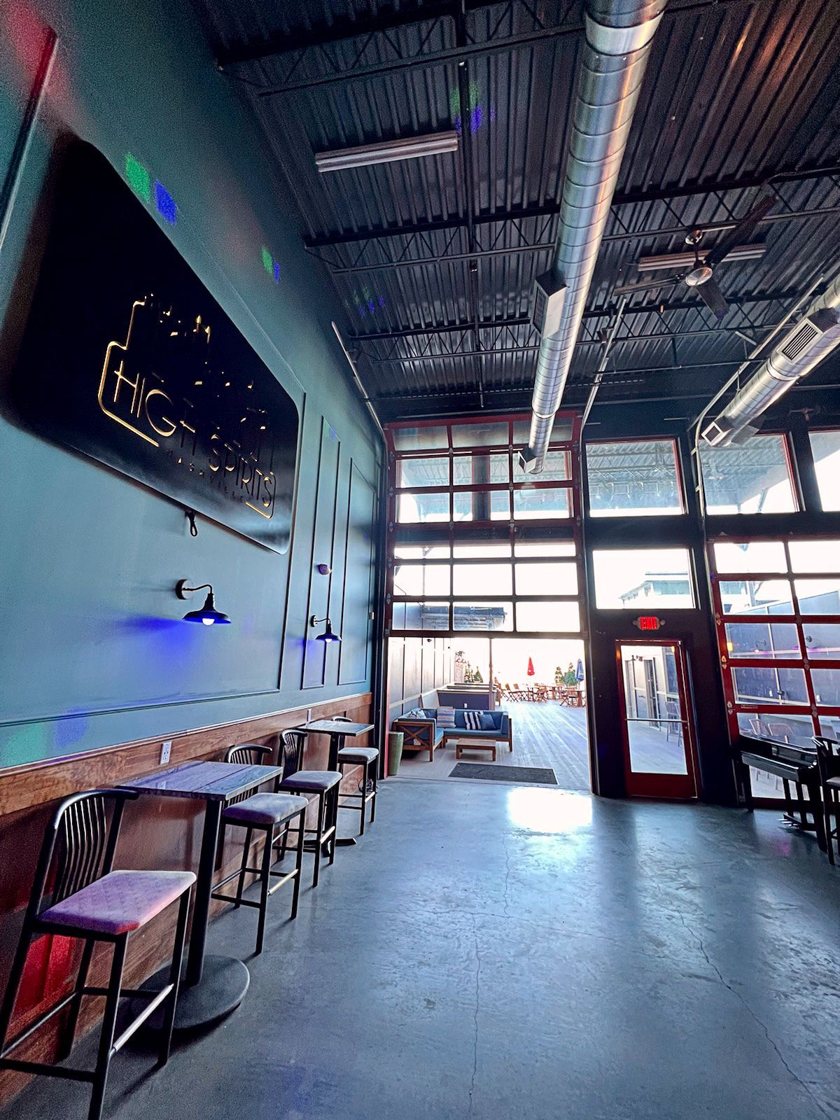 Interior of a bar with tall windows, blue wall, tables, and a neon sign.