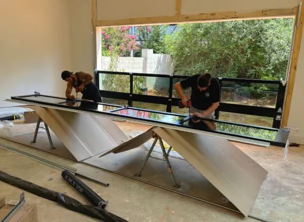 Two workers installing a long, black window panel, with a view of greenery outside.