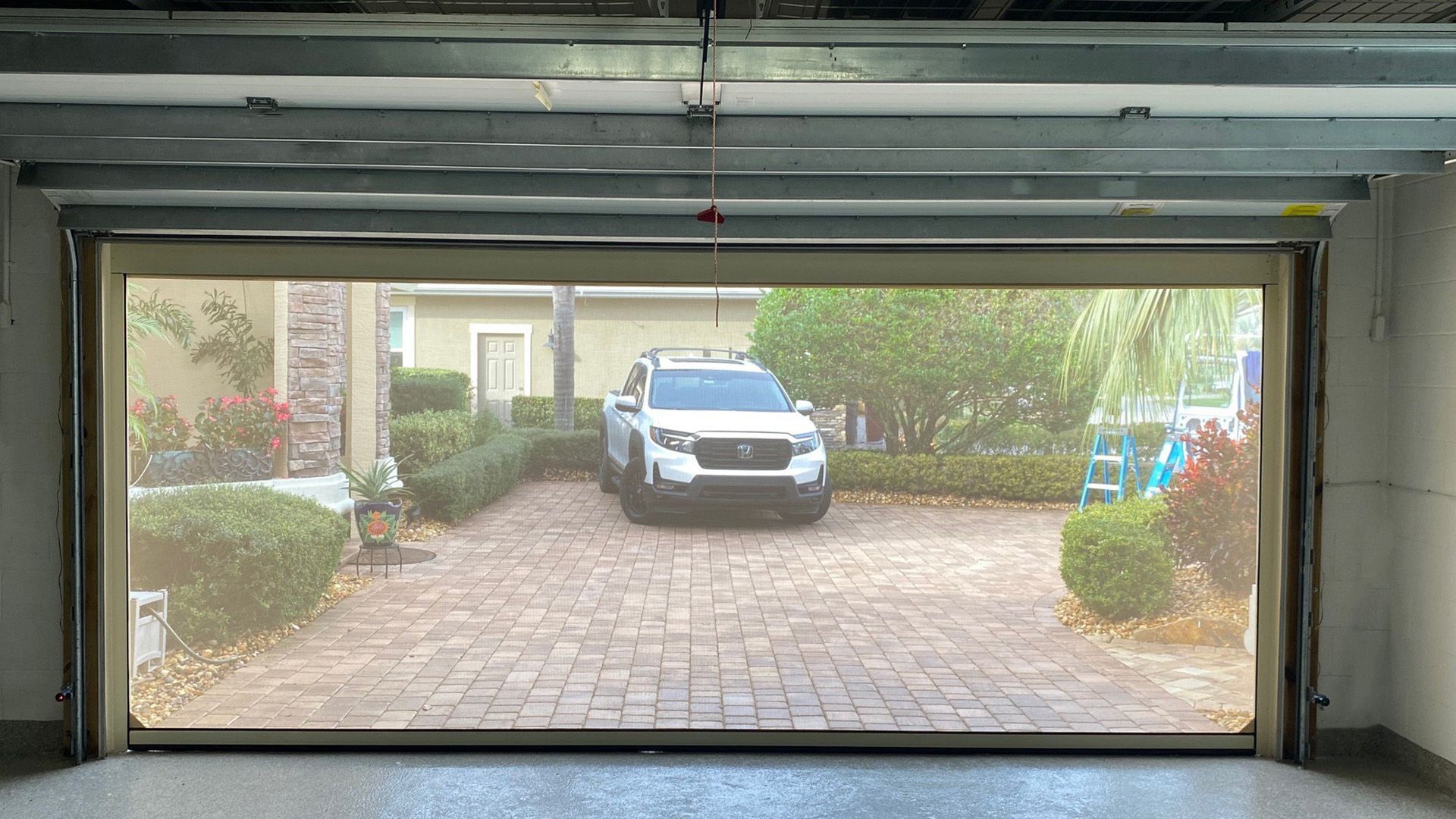 View from inside a garage with a retractable screen, closed, looking out at a white SUV parked on a paved driveway with landscaping.