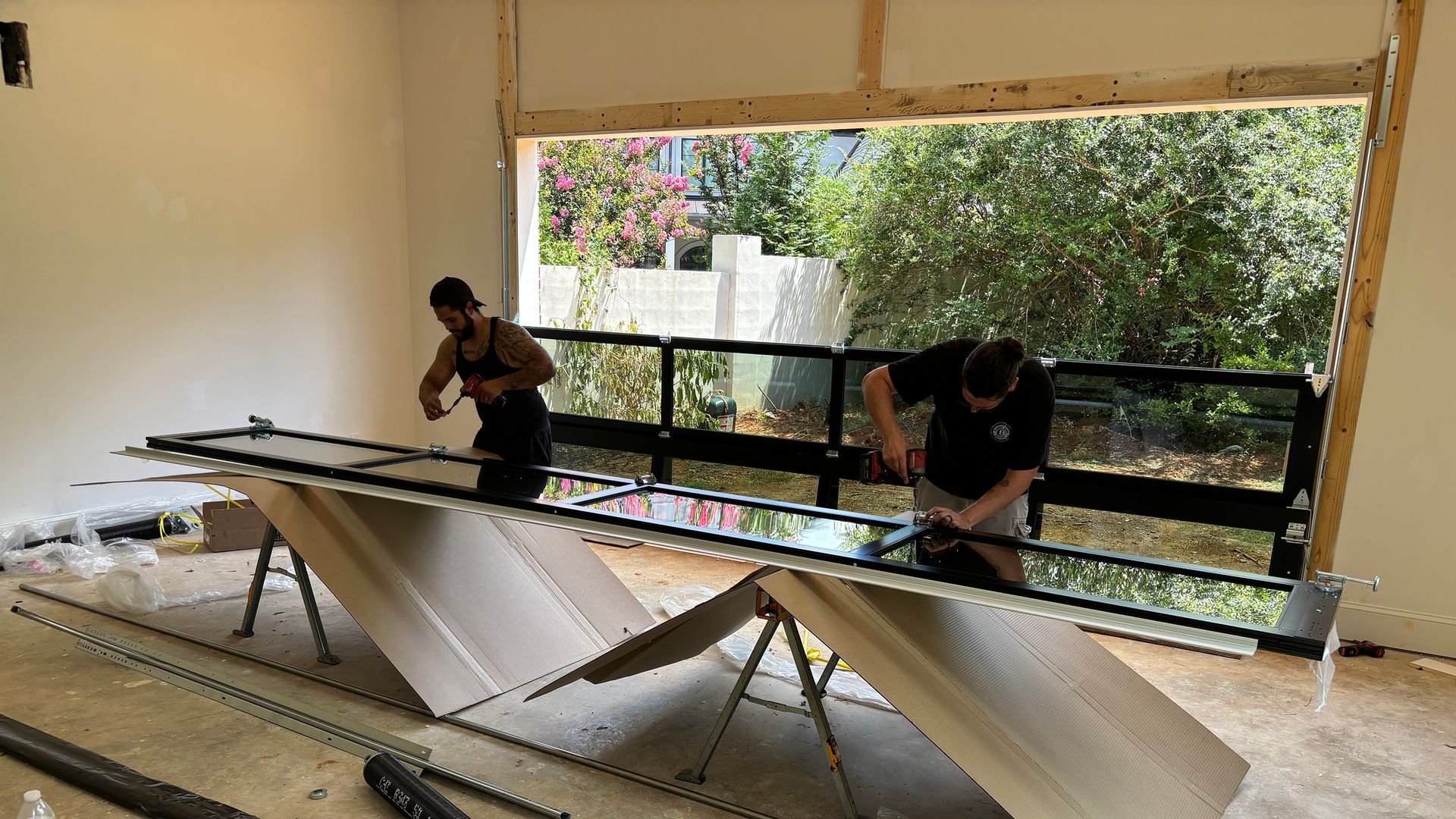 Two workers installing glass garage door panels into a frame of a building. Outdoors visible.