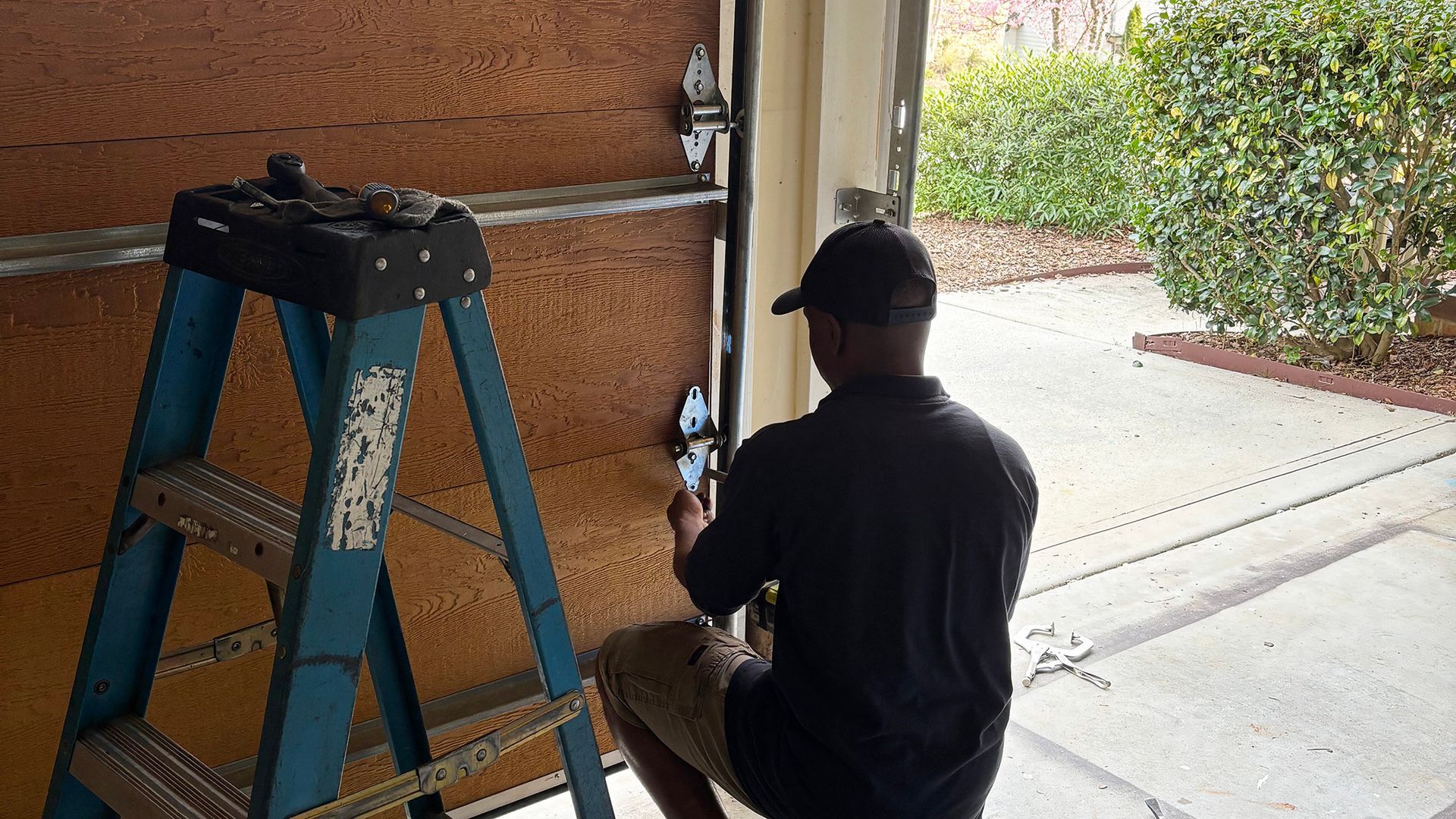 Man fixing a garage door; he's on a ladder, working near the door's hinges.