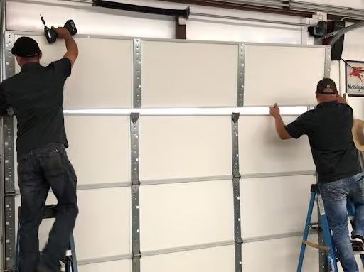 Two men installing a white garage door, using ladders and power tools inside a garage.