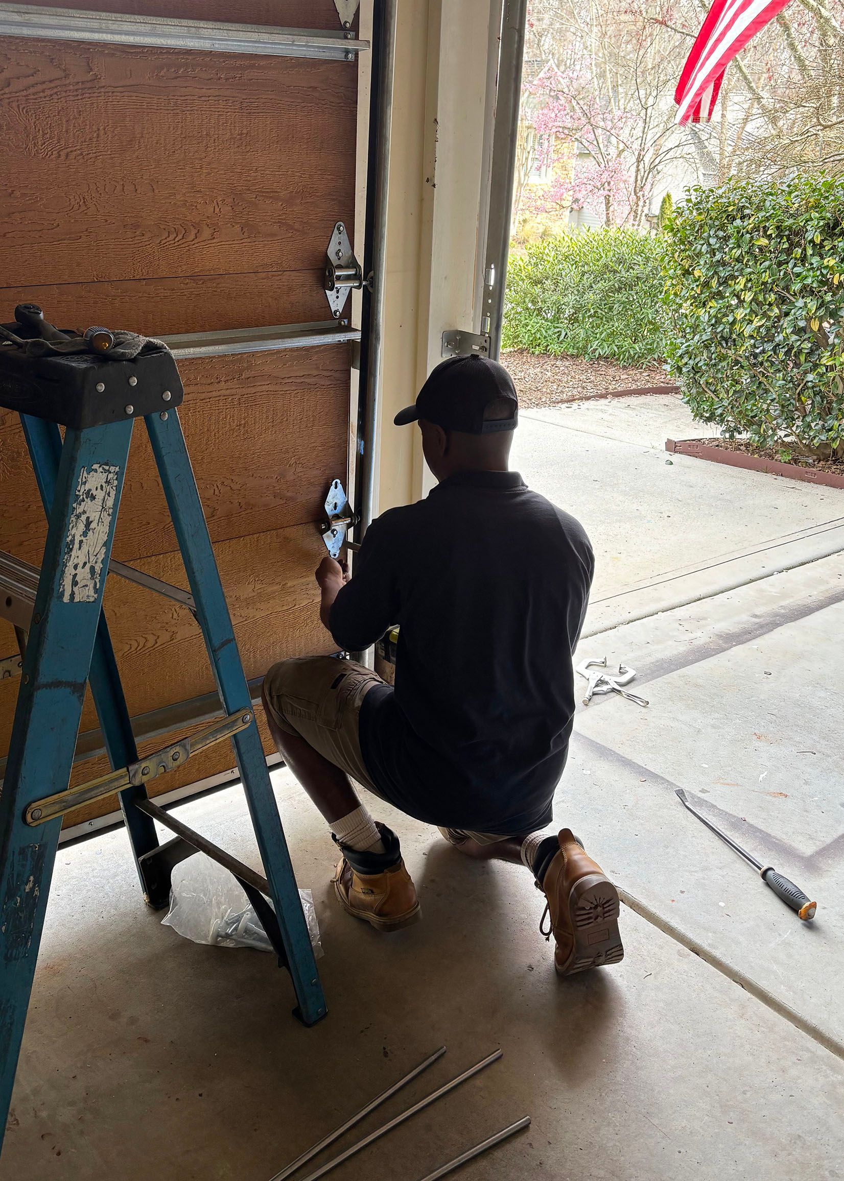 Man in black shirt and shorts repairs a garage door, using a ladder inside the garage.