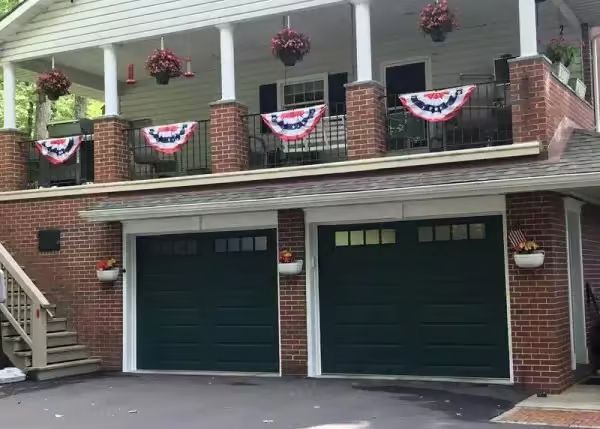 Two green garage doors beneath a brick home's porch decorated with red, white, and blue bunting and hanging flower baskets.