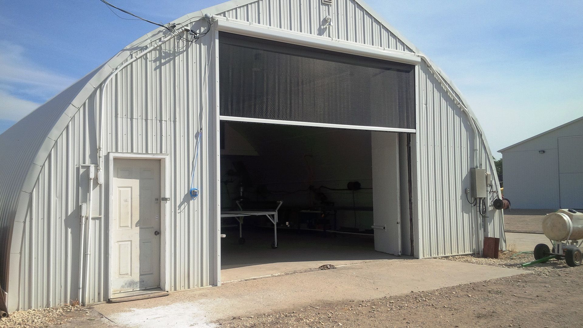 A white, arched metal Quonset-style storage building with a single door and a wide, open industrial entrance with a retractable screen.
