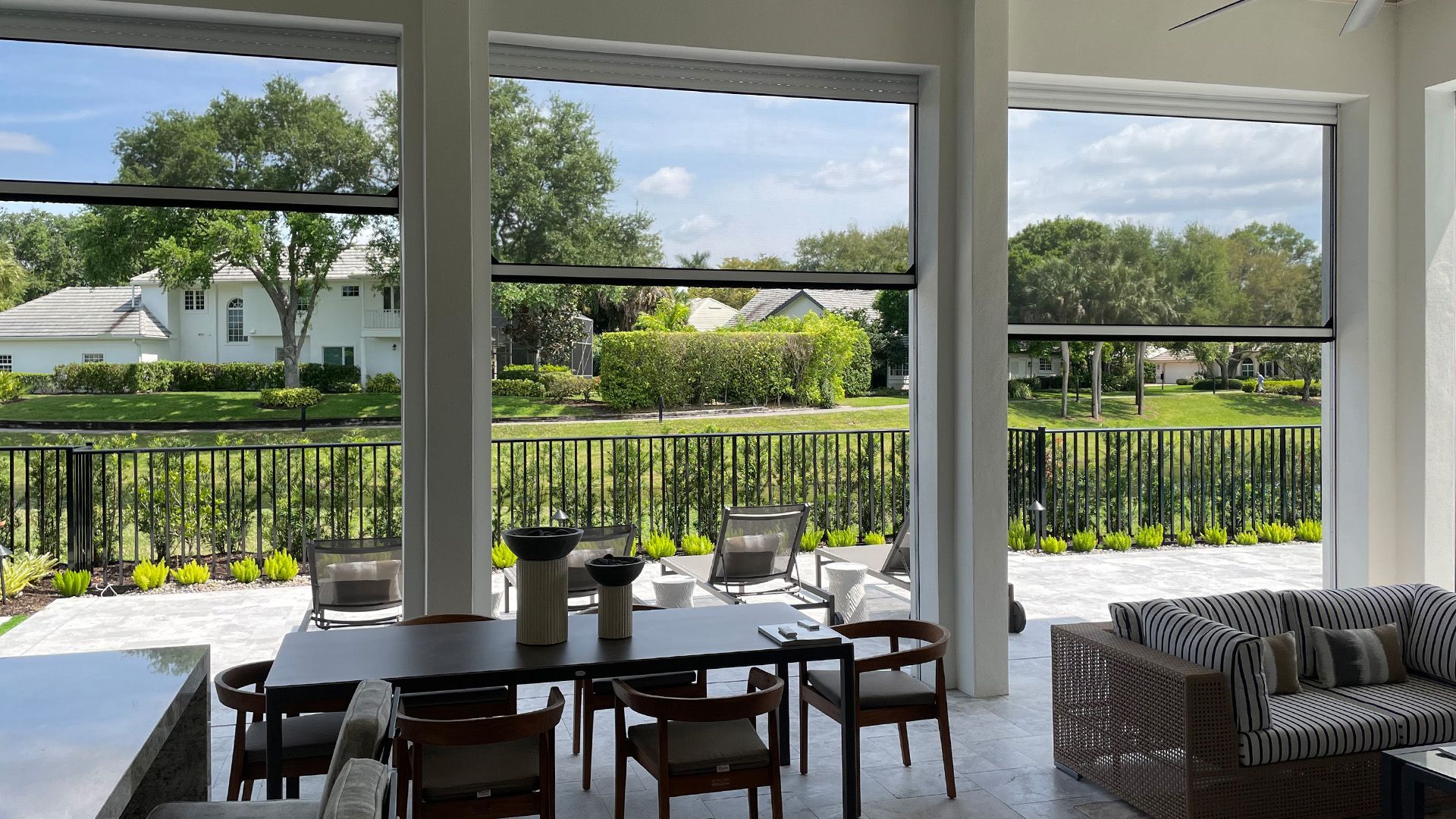A patio dining and lounge area featuring large, retractable dark screens overlooking a backyard with lush landscaping.