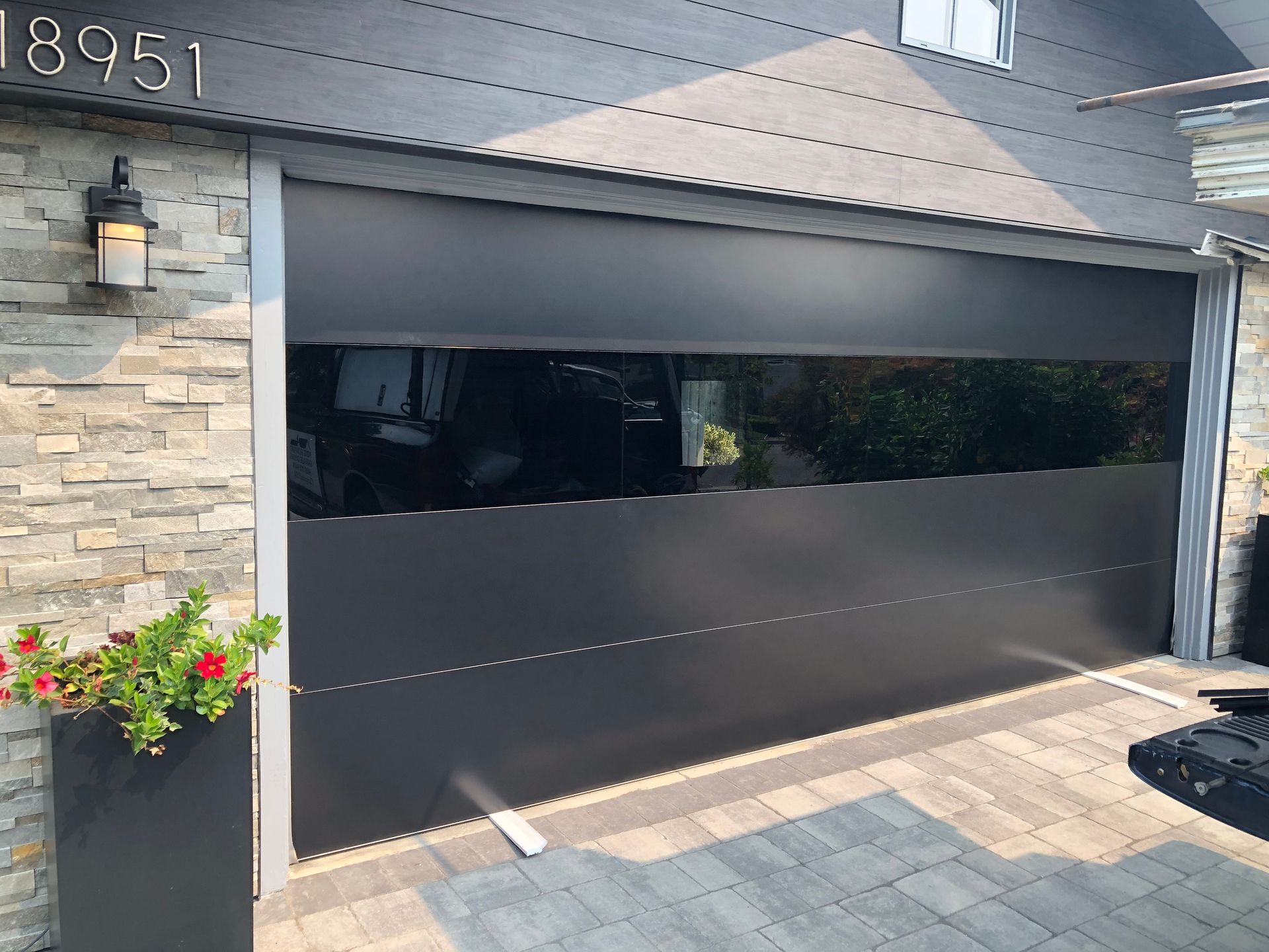 Black garage door partially open, gray trim, brick wall. House number 18951. Flower pot to the left.