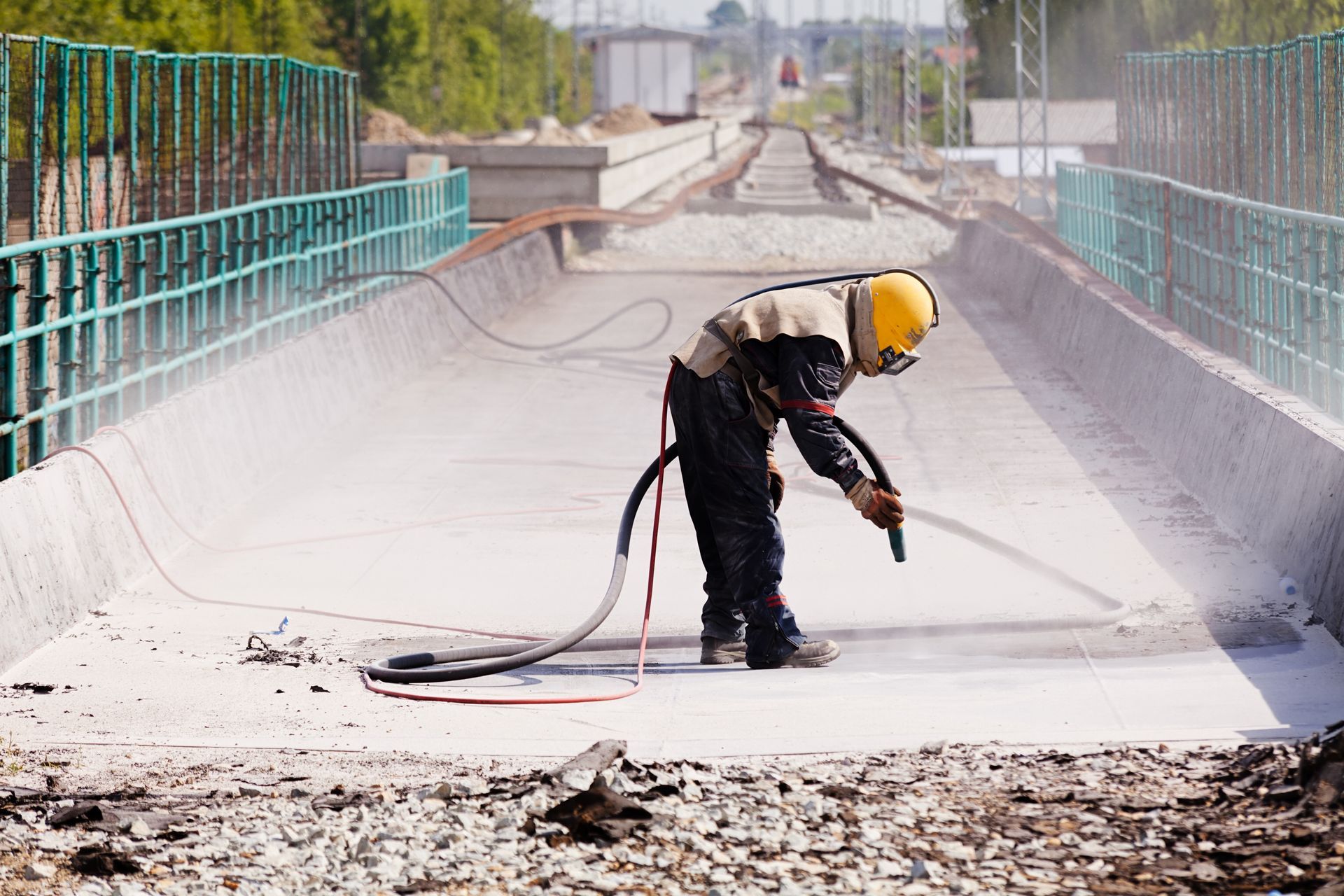 A worker in a safety helmet and vest uses an air hose to clean a section of a concrete bridge structure.