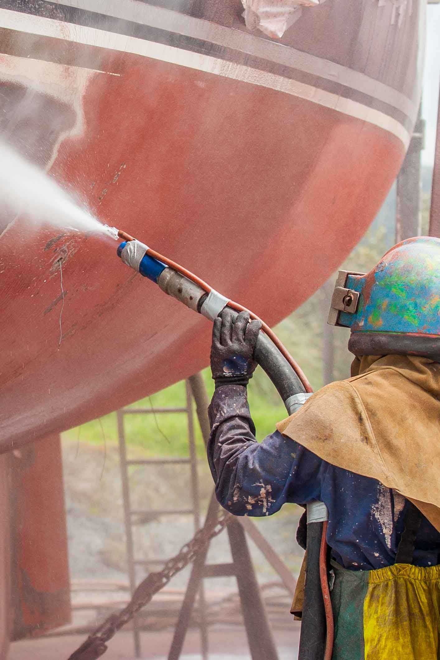 A man wearing a helmet is sandblasting a boat.