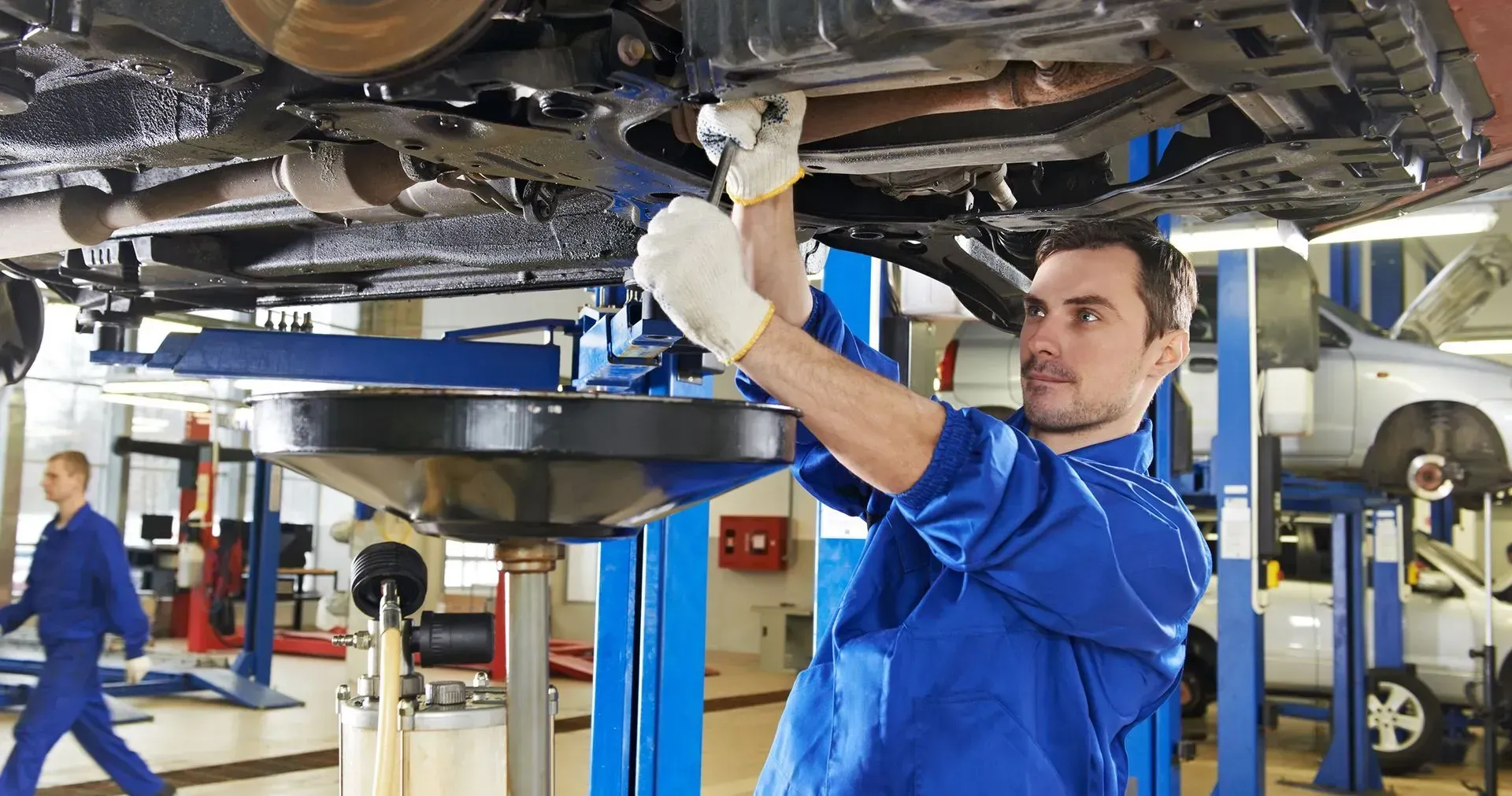 Mechanic in blue overalls changing oil under a car, oil pan below. Auto repair shop interior.