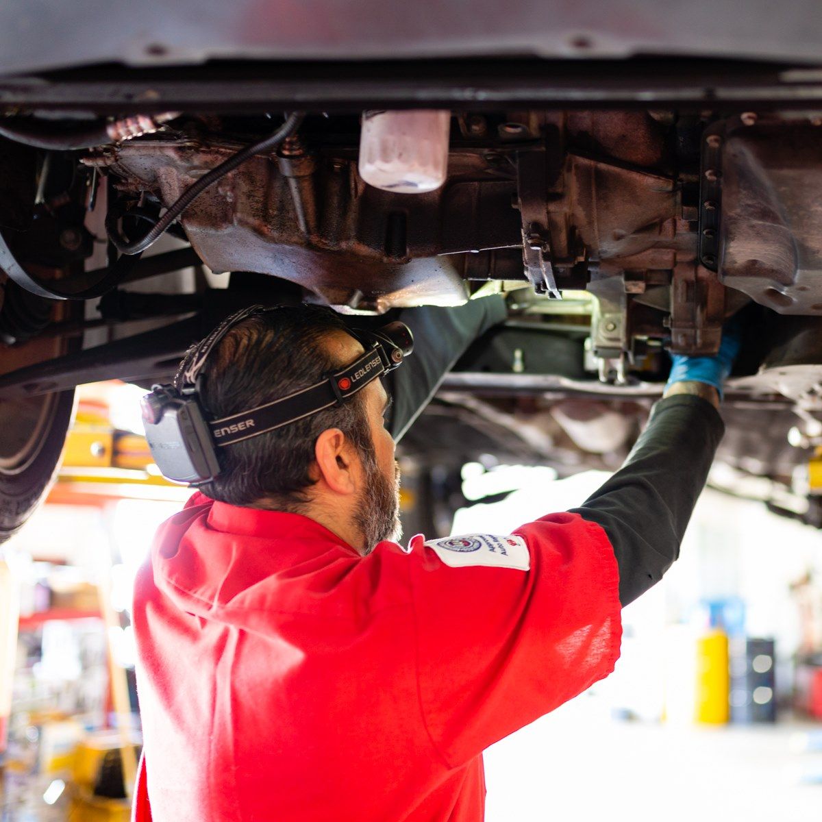 Mechanic working under a car, illuminated by a headlamp. Wears a red shirt and gloves.