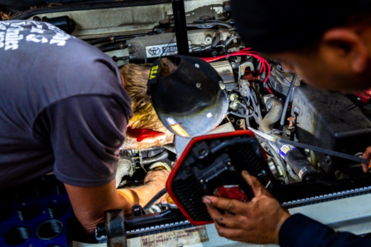 Two people working on a car engine, one using a light, and the other reaching in.
