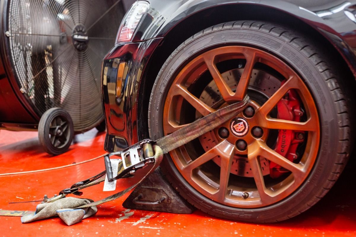 Black car's brown wheel strapped down with red brake calipers. Red floor, large fan in the background.