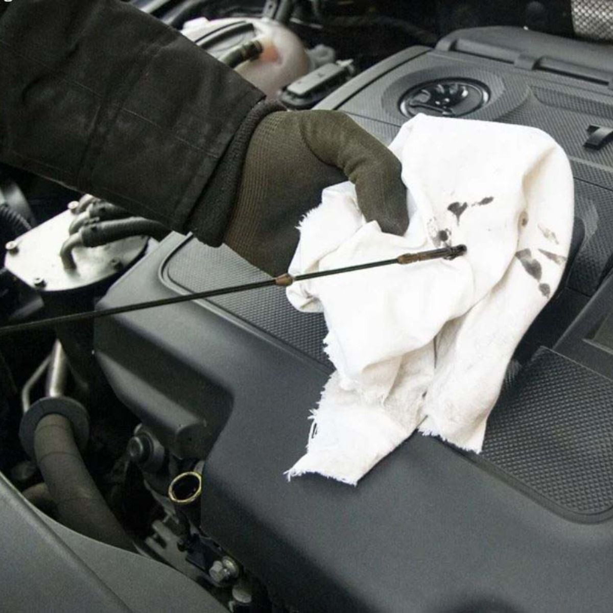 Gloved hand wiping a car's oil dipstick with a cloth, next to a black engine.