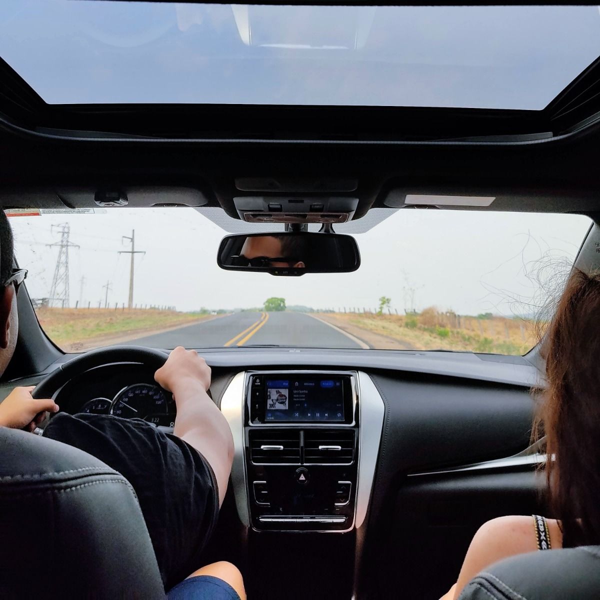 Inside a car, driving on a road with a sunroof open. Person driving with passenger. Clear sky.