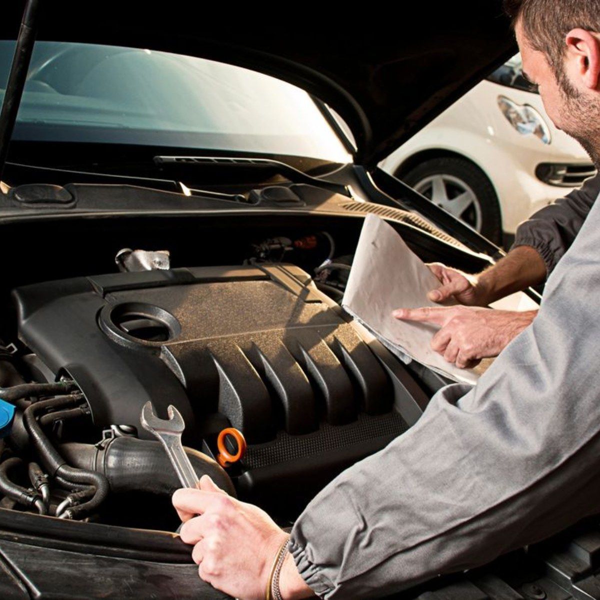 Mechanic examining car engine, holding wrench, consulting a paper. Another car in the background.