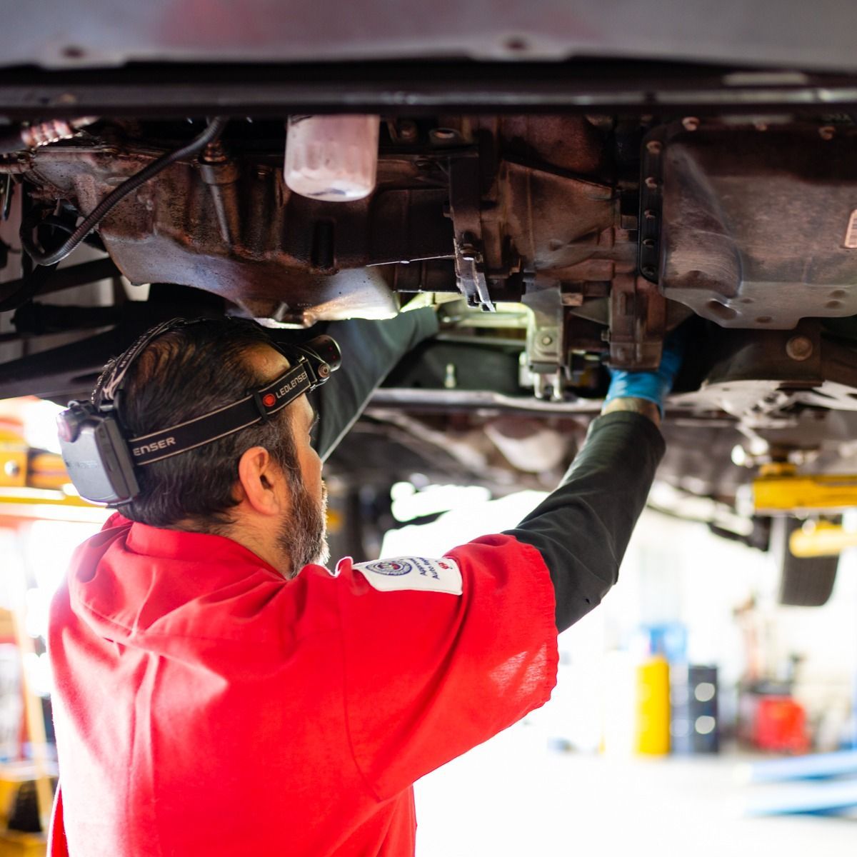 Mechanic in red shirt working under a car, illuminated by a headlamp.