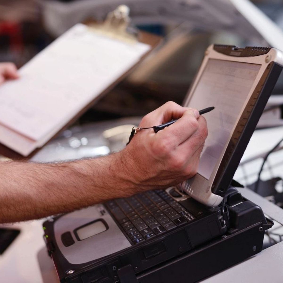 Person using a stylus to write on a laptop, holding a clipboard in a garage setting.