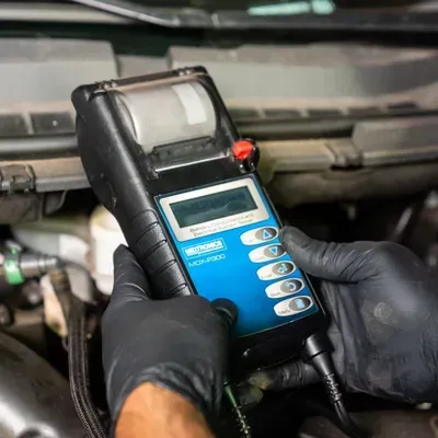 Hands in black gloves hold a blue and black diagnostic tool near a car engine.