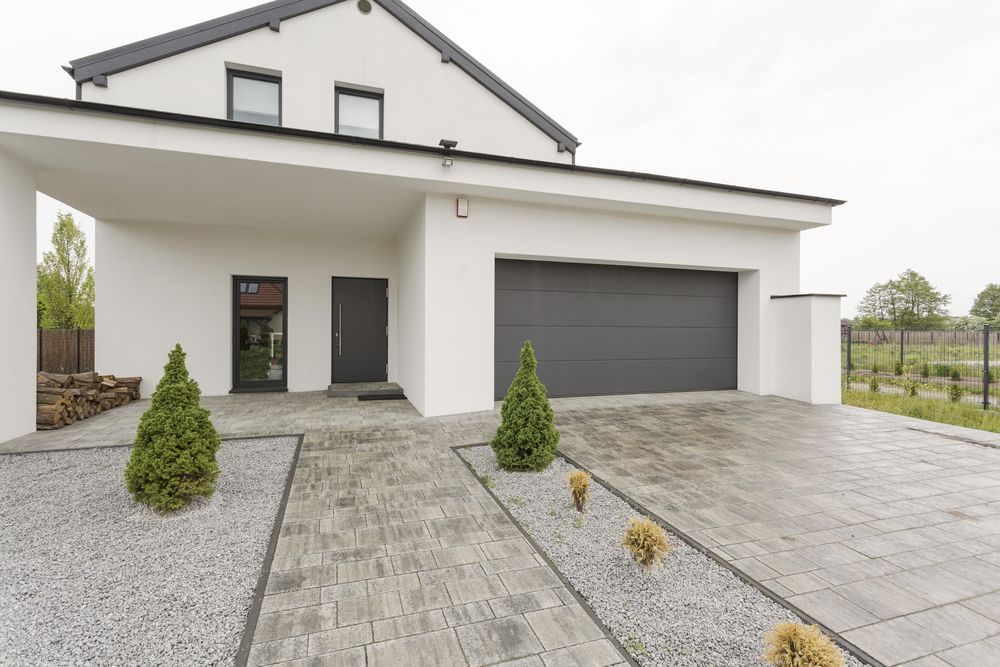 Modern white two-story house with a grey garage, dark door, and landscaped paver driveway with small evergreen trees. — On The Grind In Moreton Bay, QLD