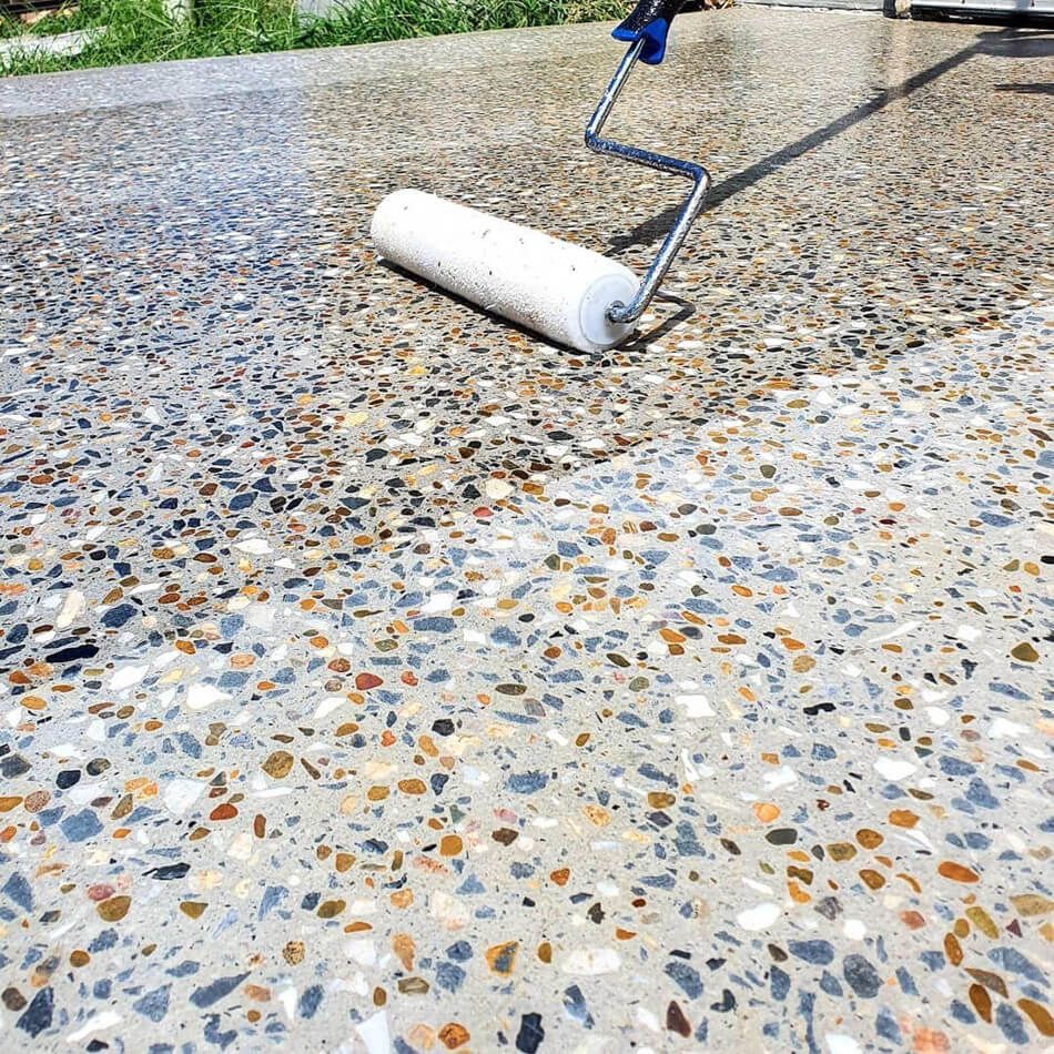 A paint roller applying a clear sealer to a concrete surface with exposed multi-colored aggregate stones. — On The Grind In Moreton Bay, QLD