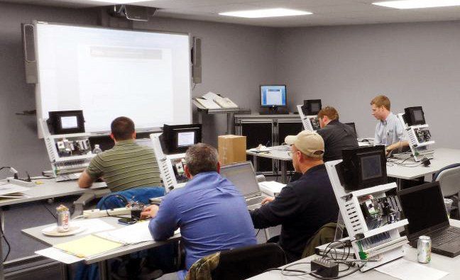 Um grupo de homens está sentado em frente a computadores em uma sala de aula.