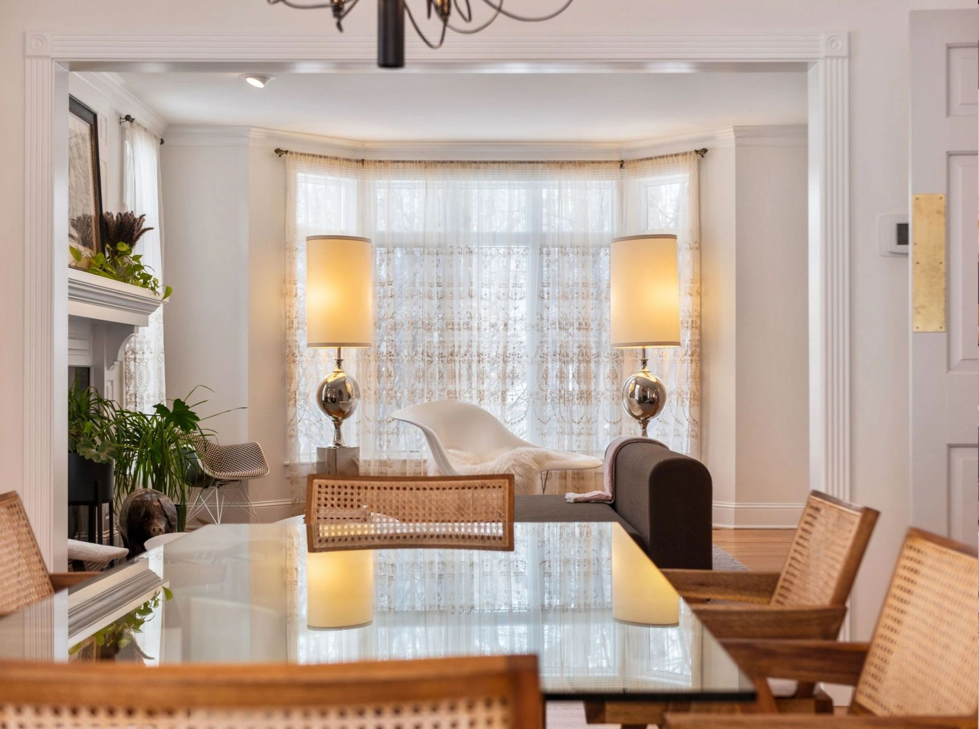 Dining room with glass table, cane chairs, and view of a sunny living area with lamps and a window seat.