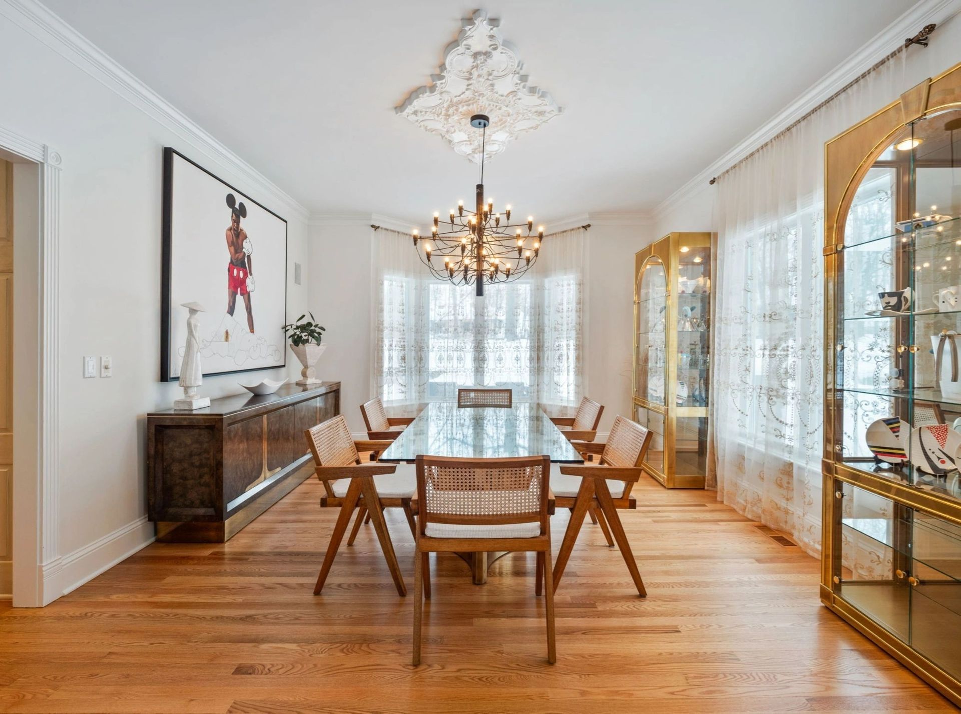 Dining room with wood floor, glass table, chandelier, art, and gold display cabinets.