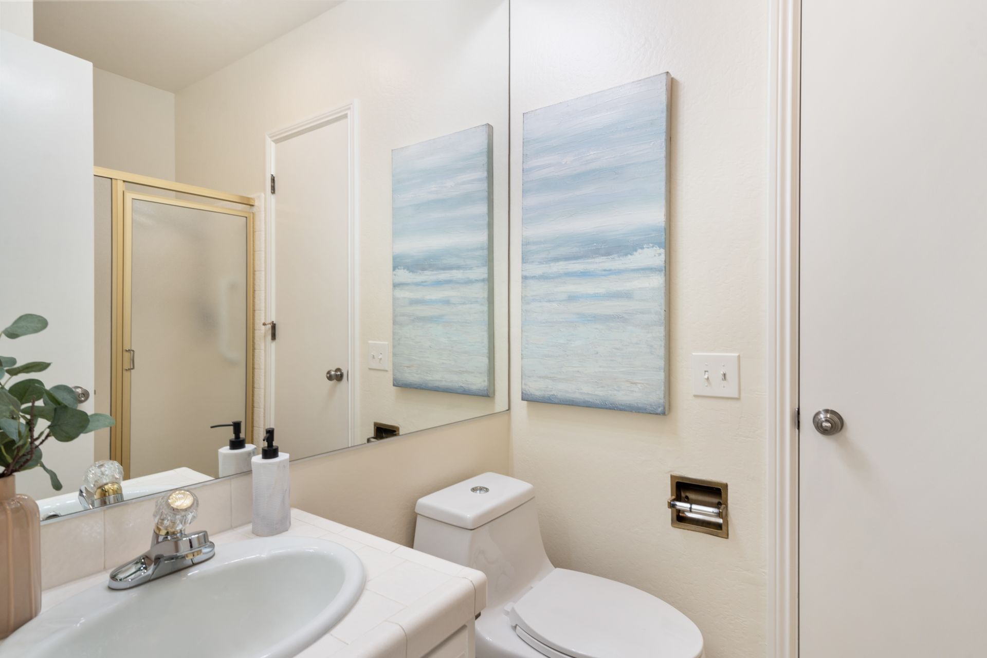 A bathroom with a sink , toilet , mirror and two paintings on the wall.