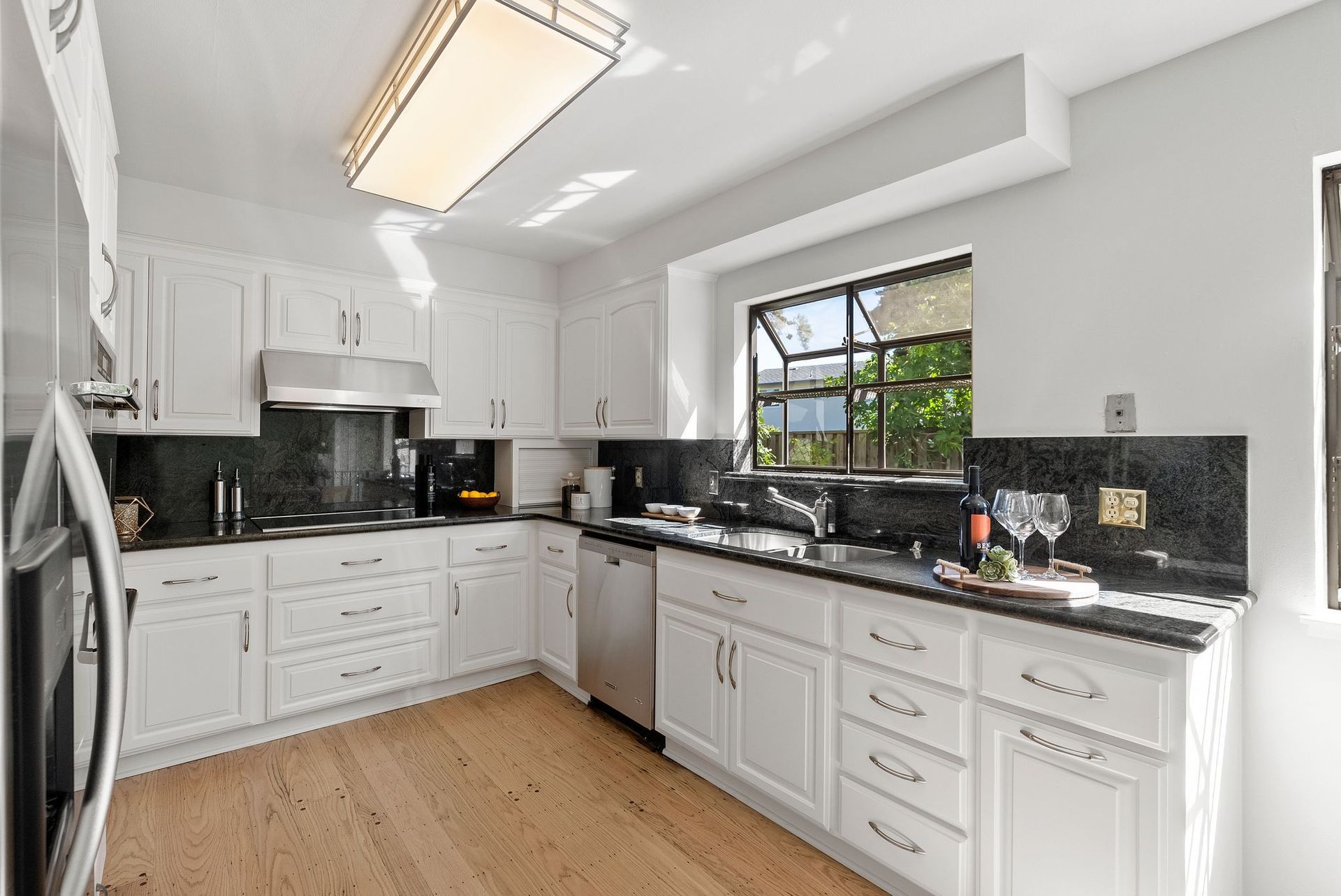 A kitchen with white cabinets , black counter tops , stainless steel appliances and a large window.