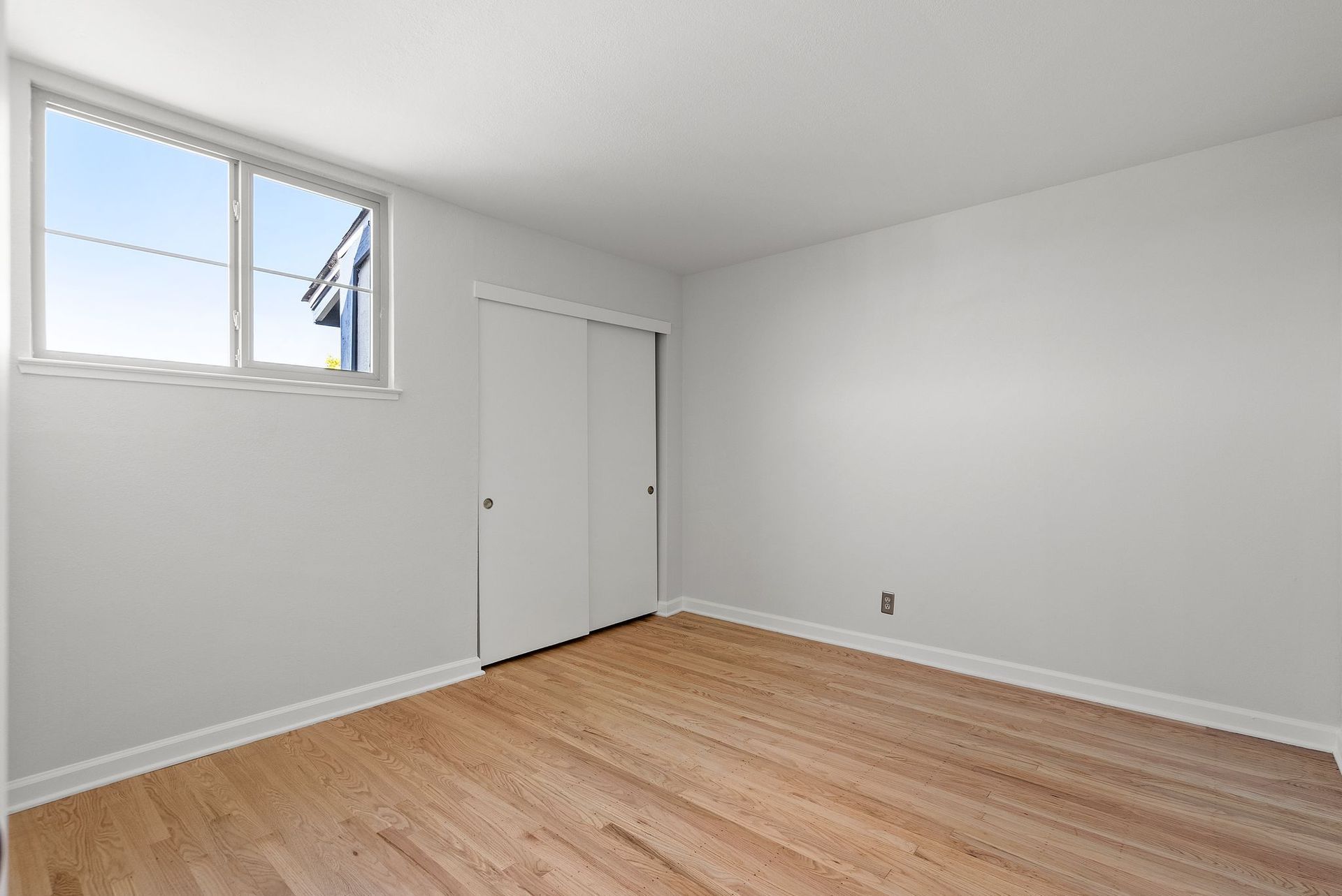 An empty bedroom with hardwood floors , white walls and a window.