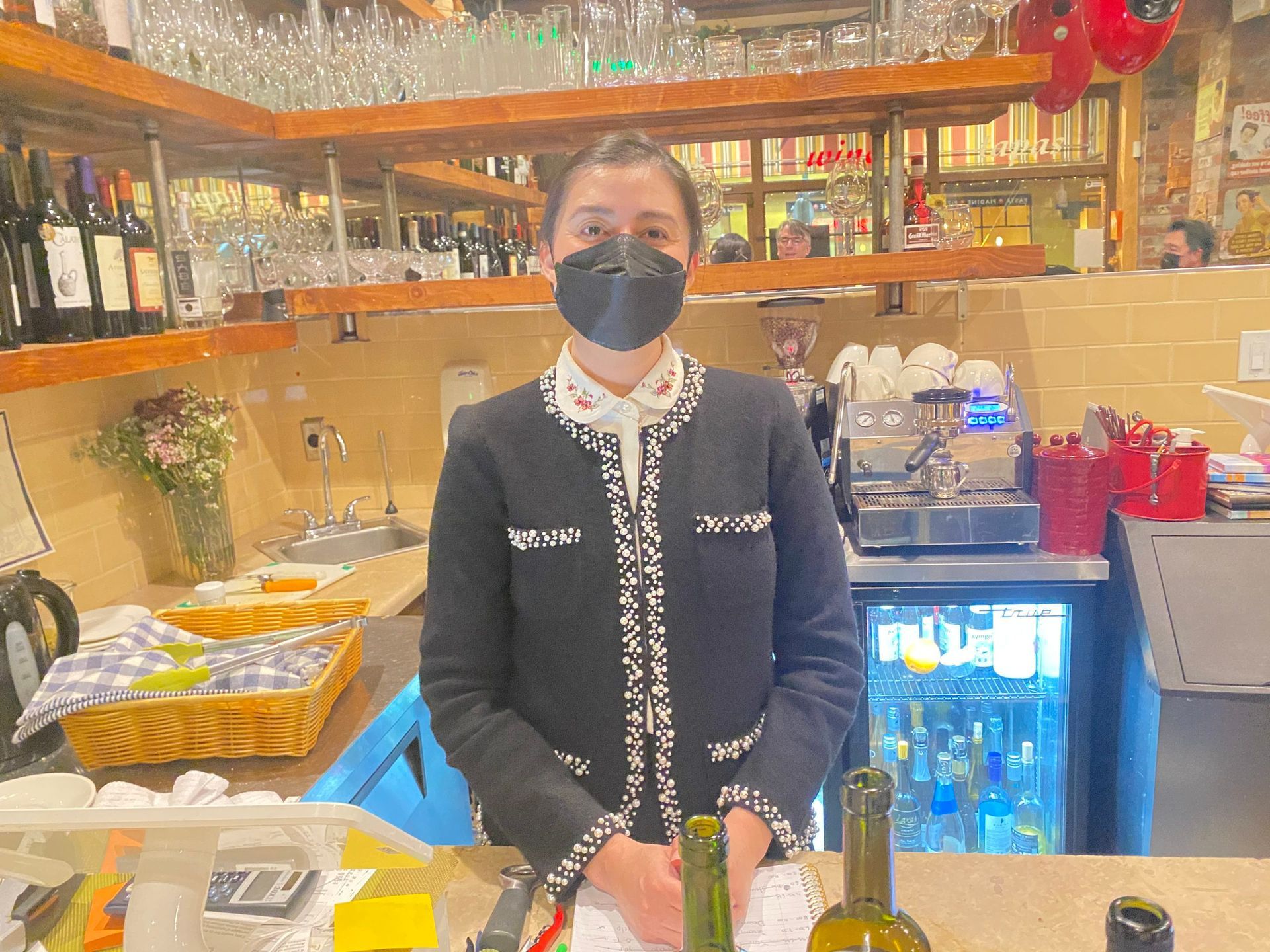 A woman wearing a mask is standing behind a bar holding bottles of wine.